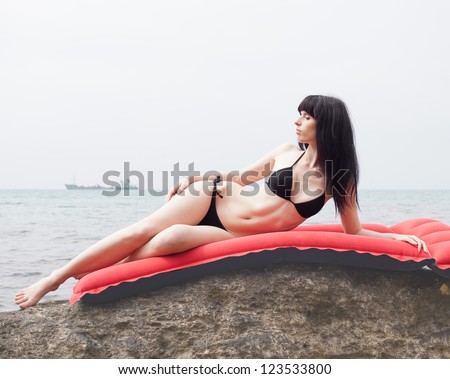 Young woman on wild rocky seashore. Attractive brunette lies on side on swimming mattress