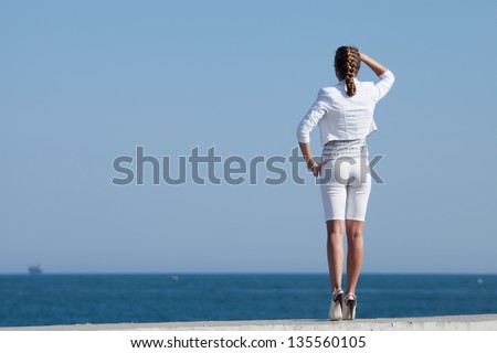 Young woman in white at the sea. Attractive woman in white stands on seafront looking into distance. Rear view