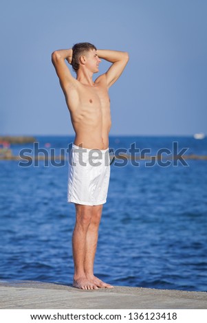 Young man in white shorts at the sea. Guy with arms raised standing on pier looking into distance