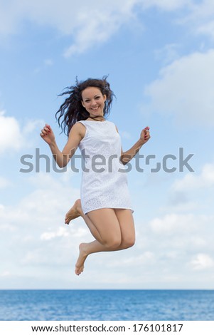 Girl in white at the sea. Young woman jumping outdoors looking at camera smiling