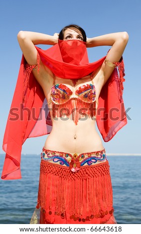 Attractive brunette in red costume for oriental dancing on the seashore. Young woman is hiding her face behind red veil
