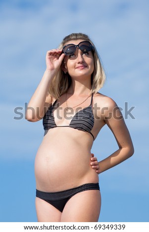 Pregnant woman in swimwear outdoors. Attractive expectant mother in bikini looking from under sunglasses at camera smiling