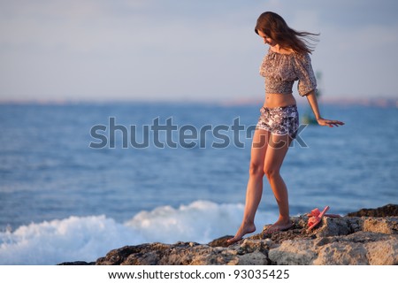 Lady at the sea. Attractive young woman in shorts and blouse on background of sea. Barefoot girl walking along the rocky beach