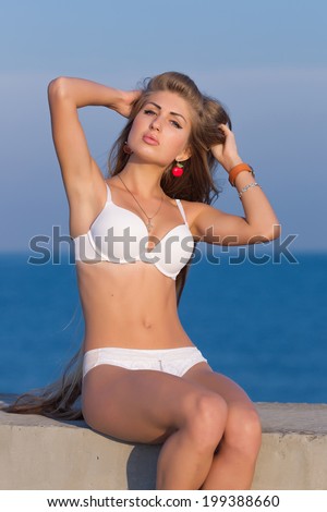 Long-haired girl on the beach. Young woman in white lingerie sits on concrete wall corrects own hair looks at camera