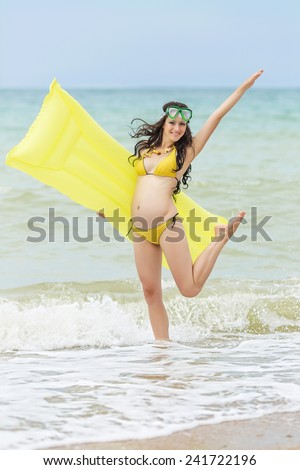 Expectant mother with pool raft and diving mask playing at the sea. Pregnant woman in yellow bikini on the beach