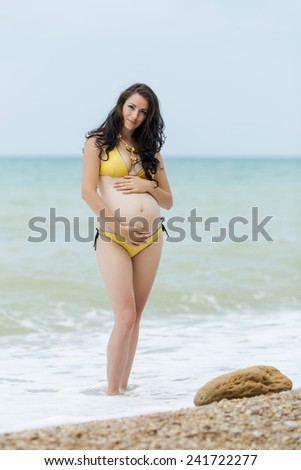 Expectant mother embracing own tummy looking at camera. Pregnant woman in yellow bikini posing on the beach