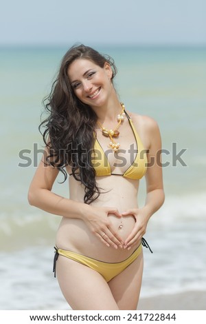 Expectant mother in yellow bikini at the sea. Pregnant woman posing a heart with her fingers on her naked belly with piercing in belly button