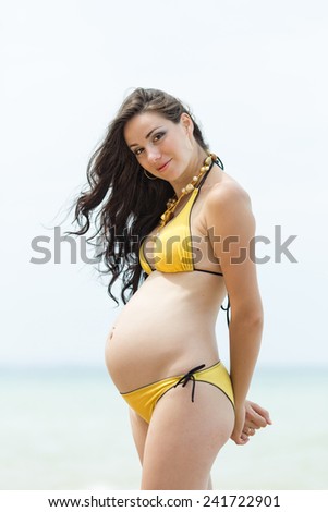 Expectant mother with hands behind his back on the beach. Pregnant woman in yellow bikini at the sea
