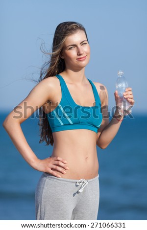 Athletic girl on open air. Attractive young woman in sports bra with plastic bottle in her hand looking at camera smiling