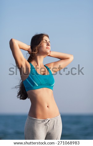Athletic girl on beach. Young long-haired woman standing against of sea with hands on head. She sunbathes with eyes closed