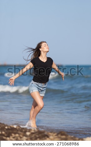 Attractive girl at the sea. Barefoot young woman in black blouse and shorts runs along seashore with arms outstretched and eyes closed