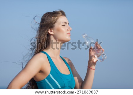Athletic girl on open air. Attractive young woman in sports bra drinks water from plastic bottle on background of sky. Waist up portrait, eyes closed, side view