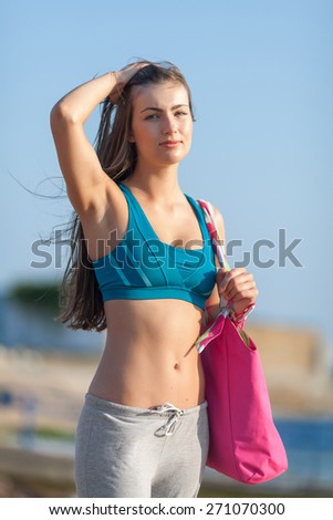 Athletic girl on open air. Young long-haired woman in sports bra with pink bag. Waist up portrait, looking at camera, one hand on head