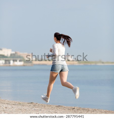Jogging. Woman in jeans shorts and grey sport uniform runs along seashore, rear view