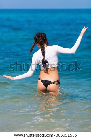 Girl at the sea. Slim brunette in white long sleeves crop top comes in water, rear view
