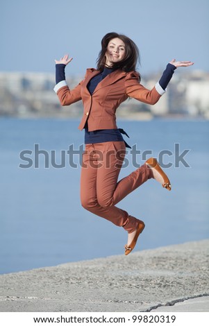 Attractive long haired brunette in brown suit on open air. Young businesswoman jumping on seafront