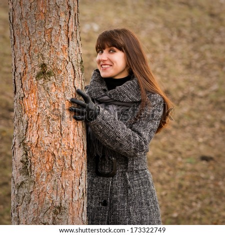 Beautiful woman in stylish winter fashion wearing a long belted knitted jumper and gloves holding the trunk of a pine tree outdoors and smiling at the camera