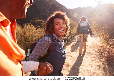 Woman Helping Man On Trail As Group Of Senior Friends Go Hiking In Countryside Together