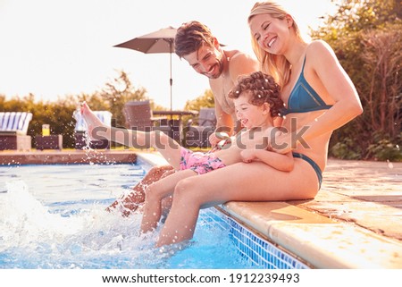 Family With Young Son Sitting On Edge Of Pool Having Fun On Summer Vacation In Outdoor Swimming Pool