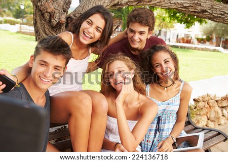 Group Of Teenagers Sitting On Bench Taking Selfie In Park