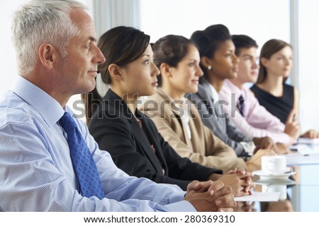 Line Of Business People Listening To Presentation Seated At Glass Boardroom Table