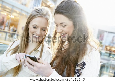 Two Female Friends Shopping In Mall Looking At Mobile Phone