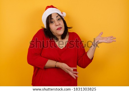 Middle aged woman wearing Christmas hatl standing against yellow background, pointing aside with both hands showing something strange and saying: I don't know what is this. Advertisement. Stok fotoğraf © 
