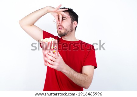 Displeased Young handsome man in red T-shirt against white background eating popcorn plugs nose as smells something stink and unpleasant, feels aversion, hates disgusting scent. Imagine de stoc © 