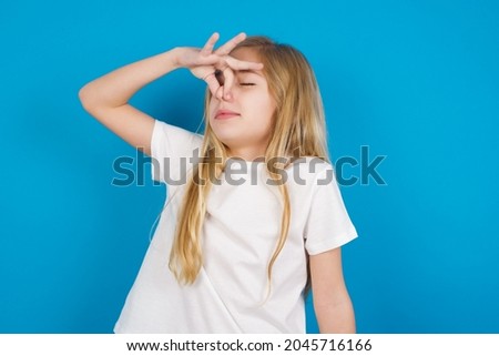 Displeased beautiful caucasian little girl wearing white T-shirt over blue background plugs nose as smells something stink and unpleasant, feels aversion, hates disgusting scent. Imagine de stoc © 
