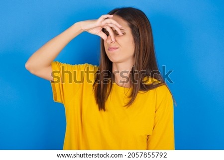 Displeased Young european brunette woman wearing yellow T-shirt on blue background plugs nose as smells something stink and unpleasant, feels aversion, hates disgusting scent. Imagine de stoc © 
