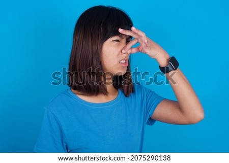 Displeased young asian woman wearing blue t-shirt against blue background plugs nose as smells something stink and unpleasant, feels aversion, hates disgusting scent. Imagine de stoc © 