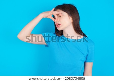 Displeased Young caucasian girl wearing blue T-shirt isolated over blue background plugs nose as smells something stink and unpleasant, feels aversion, hates disgusting scent. Imagine de stoc © 
