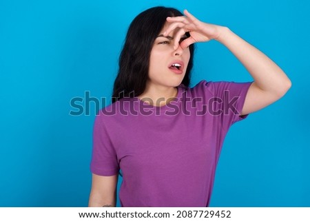 Displeased Caucasian woman wearing purple T-shirt over blue background plugs nose as smells something stink and unpleasant, feels aversion, hates disgusting scent. Imagine de stoc © 