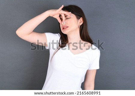 Displeased young beautiful Caucasian woman wearing white T-shirt over studio grey wall, plugs nose as smells something stink and unpleasant, feels aversion, hates disgusting scent. Imagine de stoc © 