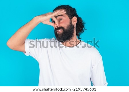 Displeased Caucasian man with beard wearing white t-shirt over blue background plugs nose as smells something stink and unpleasant, feels aversion, hates disgusting scent. Imagine de stoc © 