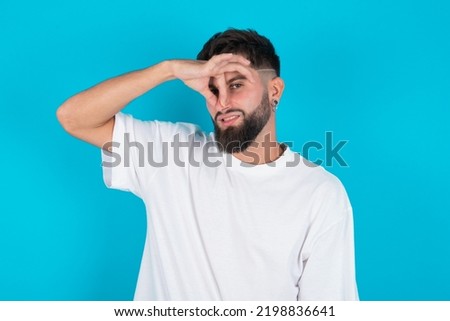 Displeased bearded caucasian man wearing white T-shirt over blue background plugs nose as smells something stink and unpleasant, feels aversion, hates disgusting scent. Imagine de stoc © 