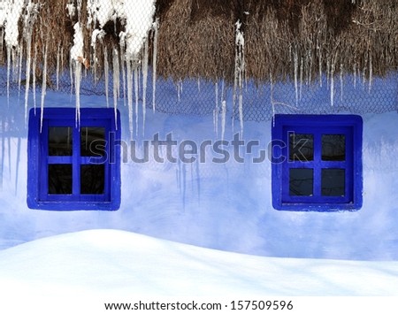 Rustic house with straw roof and blue window is covered with snow
