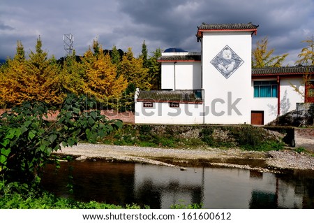SICHUAN PROVINCE, CHINA: A white Chinese house with a diamond-shaped wall mural overlooks a small river next to a grove of Gingko trees with yellow Autumnal leaves