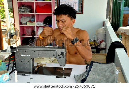 Hua Hin, Thailand - January 1, 2010:  Bare chested tailor using a Singer Sewing machine threading a needle at his roadside shop