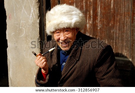 Pengzhou, China - January 25, 2007:  Elderly Chinese man wearing a fur hat and smoking a cigarette in a holder seated in front of his home on Hua Lu (Old Street)