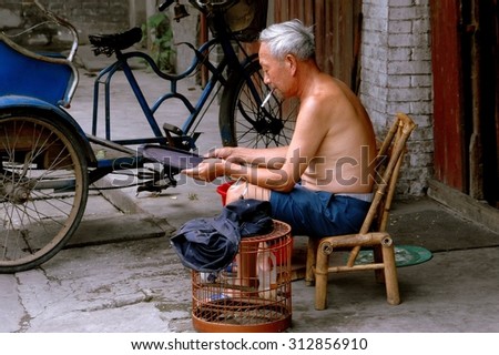 Pengzhou, China - August 27, 2006:  Shirtless Chinese man seated on a bamboo chair cleans the tray to his birdcage on Hua Lu (Old Street)