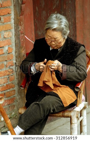 Pengzhou, China - April 13, 2006: Elderly Chinese woman cutting a piece of fabric sitting in front of her home on Hua Lu (Old Street)