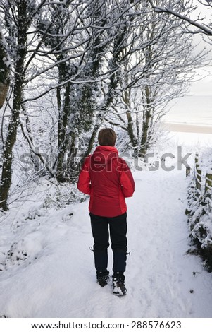 Walker in red jacket on snow covered Isle of Anglesey Coastal Path leading to the beach in winter. Red Wharf Bay (Traeth Coch), Isle of Anglesey, North Wales, UK, Britain.