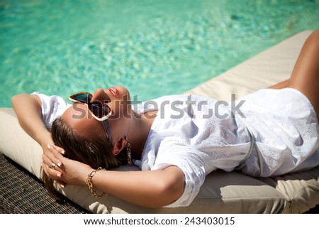 Beautiful woman holding hand behind head, lying on sun chaise longue near luxury pool side. Young woman enjoying summer vacation.