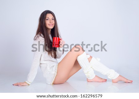 A young beautiful fashion brunette woman in white sweater no pants in getters holding a red mug of coffee smiling on a white background