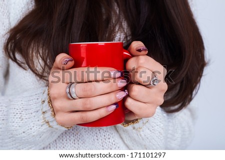 A red mug with coffee in hands of a girl with manicure, long hair and white sweater no face