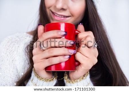 A red mug with coffee in hands of a girl with manicure, long hair and white sweater no face
