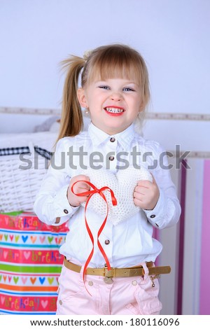 A three year old girl standing in a pink room, in white blouse and pink pants with makeup red lips and a hairstyle holding a white knitted heart