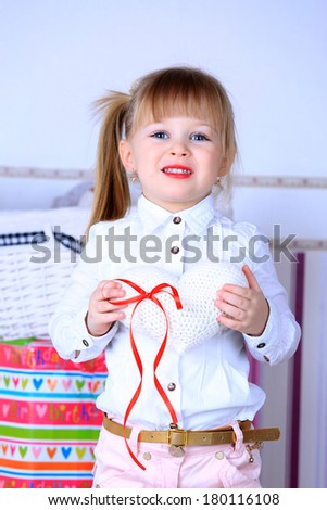 A three year old girl standing in a pink room, in white blouse and pink pants with makeup red lips and a hairstyle holding a white knitted heart