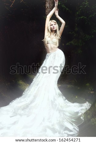 Blonde angel in long white skirt standing in dark forest